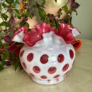 May include: A vintage white glass vase with a ruffled, fuchsia rim and fuchsia polka dots. The vase has a round body and a pedestal base. The background includes greenery and a glimpse of other decorative items.