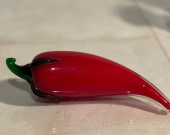 May include: A handcrafted glass chili pepper sculpture. The pepper is a vibrant red with a glossy finish, featuring a green stem and dark accents. This decorative piece is ideal for kitchen or dining room decor.