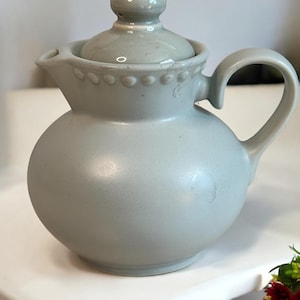 May include: A light gray ceramic teapot with a round body and a lid with a knob. The teapot has a handle and a beaded trim around the top.