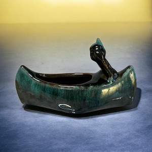 May include: A green and brown ceramic canoe-shaped planter with a bird head on the bow. The planter has a glazed finish and is perfect for displaying small plants.