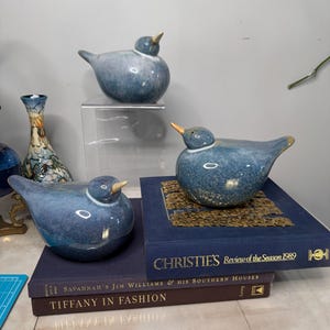 May include: Three blue ceramic bird figurines on a stack of books. The books are titled "Christie's Review of the Season 1989", "Savannah's Jim Williams & His Southern Houses", and "Tiffany in Fashion".
