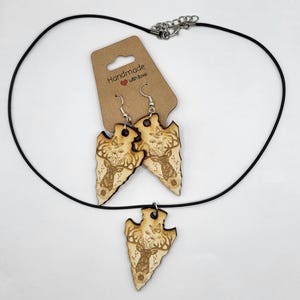 May include: A wooden necklace and earrings set featuring a deer head design with butterflies. The necklace has a black cord and the earrings have silver hooks. The set is handmade with love.