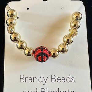 Op de afbeelding: Een gouden kralenarmband met een centrale rode en zwarte kristallen kraal. De armband heeft ook kleinere kristallen kralen en wordt gepresenteerd op een witte kaart met de tekst "Brandy Beads and Blankets".