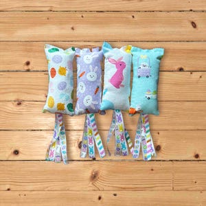 May include: Four Easter-themed bunny-shaped cat toys with colorful ribbon tails. The toys are made of fabric and feature different bunny designs, including a bunny with carrots, a bunny with eggs, a bunny with a wagon, and a pink bunny.