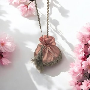 May include: A dusky pink drawstring pouch with a decorative tassel and a braided cord. The pouch has a gold trim and is suspended against a white backdrop with pink cherry blossoms.