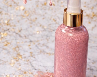 May include: A pink glitter spray bottle with a gold cap and clear top. Pink glitter drips from above, and a pool of glitter sits below the bottle. The background is a marble-like surface with gold flecks.