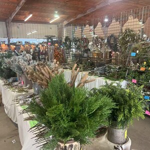 May include: A variety of greenery and branches are displayed on tables and shelves in a market setting. The greenery includes cedar, pine, and other evergreen varieties. The branches are mostly brown and have a natural, rustic look.