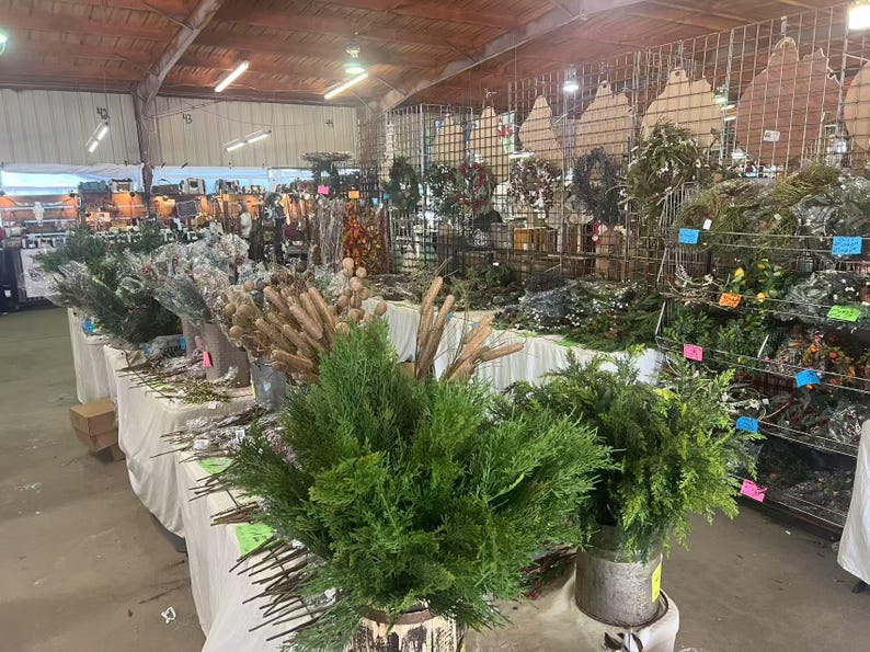 May include: A variety of greenery and branches are displayed on tables and shelves in a market setting. The greenery includes cedar, pine, and other evergreen varieties. The branches are mostly brown and have a natural, rustic look.