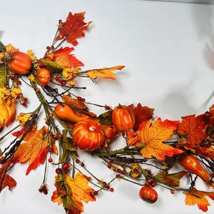 May include: A fall-themed garland featuring orange pumpkins, red berries, and colorful autumn leaves in shades of orange, yellow, and red. The garland is arranged on a white background, creating a festive and decorative display.