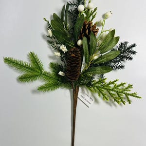 May include: A green and brown artificial floral spray with pine cones, white berries, and green leaves. The spray has a brown stem and a white tag that reads "ITEM NO. BB478 MADE IN CHINA."