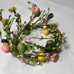 May include: A green and pink Easter wreath with faux eggs and flowers. The wreath is made of artificial greenery and has a variety of pastel-colored eggs and flowers.
