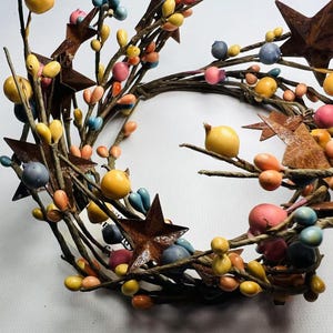 May include: A rustic wreath made of twigs with colorful berries and rusty metal stars. The wreath is decorated with pink, yellow, blue, and orange berries.