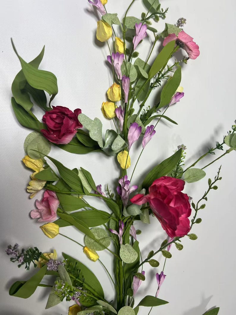 May include: A floral garland with pink, red, and yellow flowers, green leaves, and purple accents. The garland is perfect for adding a touch of spring to your home decor.