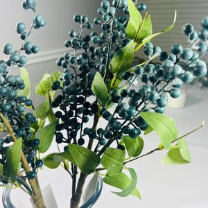 May include: A bouquet of artificial green berry sprigs with small, dark green berries arranged in a clear glass vase.
