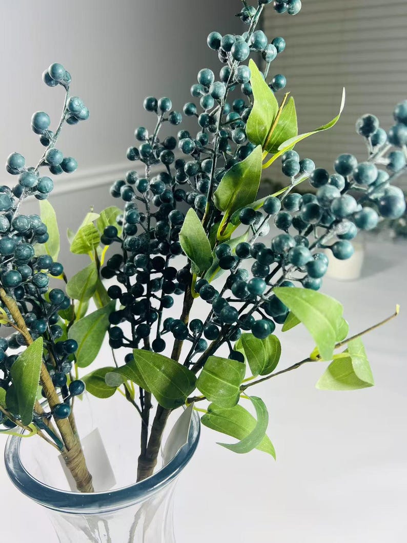 May include: A bouquet of artificial green berry sprigs with small, dark green berries arranged in a clear glass vase.