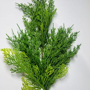 May include: A vibrant green artificial cedar branch with a brown stem. The branch features detailed, textured foliage in varying shades of green, with a lighter, yellowish-green hue at the tips. A small white tag is attached to the base of the branch.