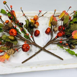 May include: Two sprigs of fall-themed artificial greenery with brown stems, green leaves, and small orange pumpkins. The sprigs are arranged on a white surface with red berries and brown pinecones.