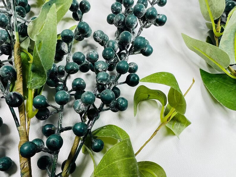 May include: A sprig of faux greenery with dark green berries and leaves. The berries have a frosted finish.