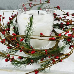 May include: A white candle in a clear glass jar surrounded by a green and red berry garland. The garland is made of artificial pine sprigs and red berries.