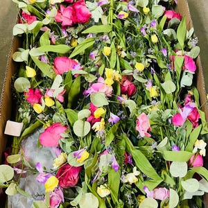 May include: A box filled with a variety of artificial flowers and greenery, including pink roses, yellow flowers, and green leaves.