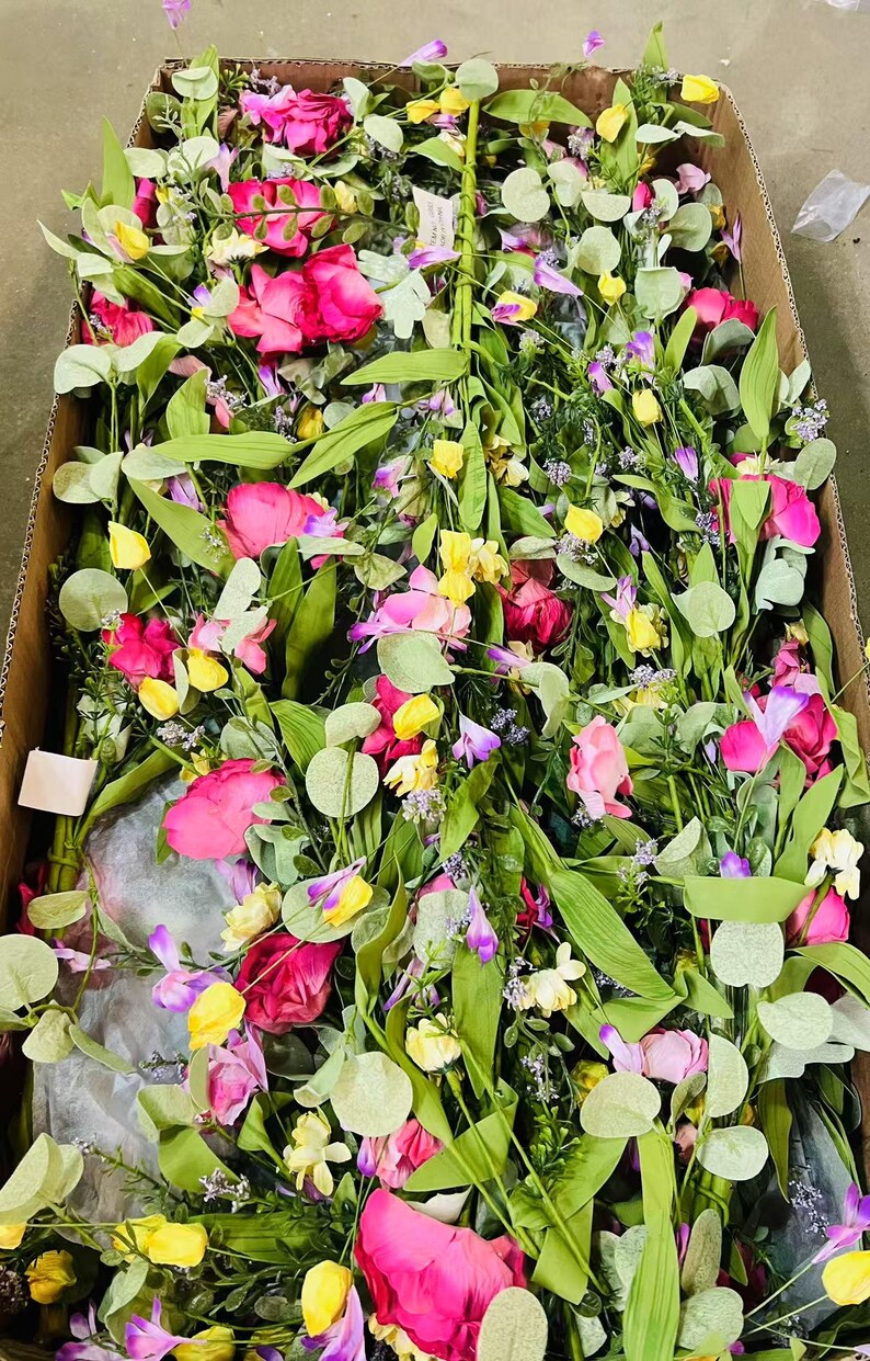May include: A box filled with a variety of artificial flowers and greenery, including pink roses, yellow flowers, and green leaves.