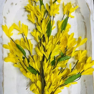 May include: A bunch of bright yellow artificial flowers with green leaves. The flowers are arranged in a bouquet and are displayed on a white background.