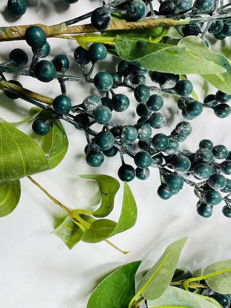May include: A sprig of faux greenery with dark green berries and leaves. The berries have a frosted look.