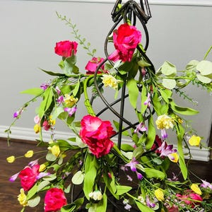 May include: A metal star-shaped stand with a spiral design is decorated with pink, yellow, and purple artificial flowers. The stand is surrounded by green leaves.