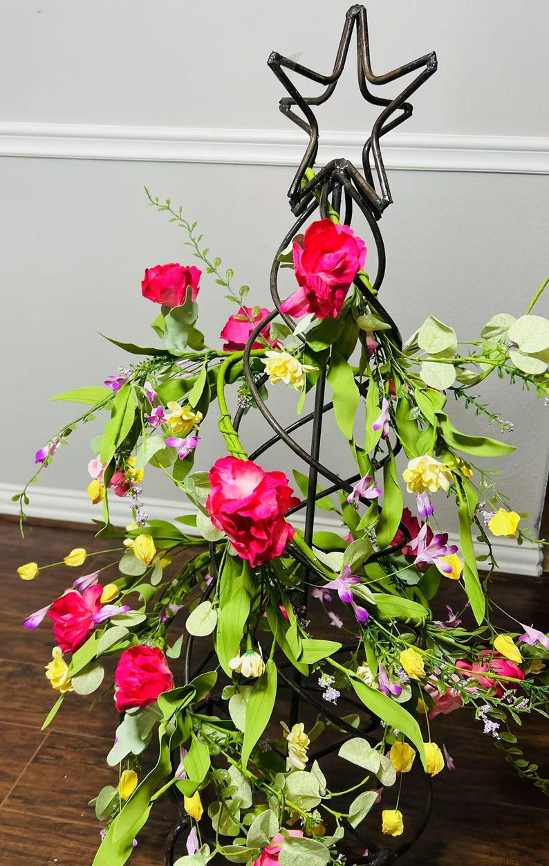 May include: A metal star-shaped stand with a spiral design is decorated with pink, yellow, and purple artificial flowers. The stand is surrounded by green leaves.