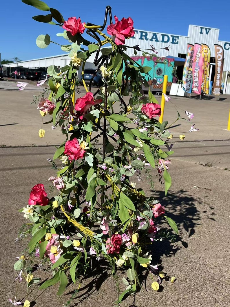 May include: A metal floral stand with a star-shaped base is decorated with pink and white flowers and green leaves. The stand is in front of a building with the word "TRADE" on it.