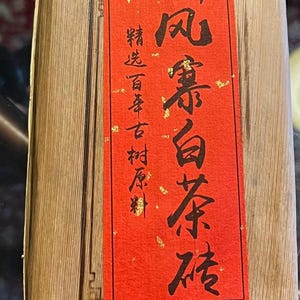 May include: A rectangular package wrapped in light brown paper, secured with a thin wire. A red label with black Chinese characters and gold accents is affixed to the front. The label features text.