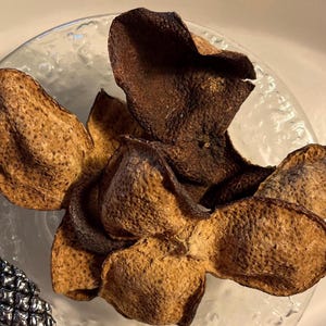 Può includere: Primo piano di scorze d'arancia essiccate disposte su un piatto di vetro trasparente e strutturato. Le scorze sono marroni e beige, con una superficie ruvida. La disposizione suggerisce un uso culinario o decorativo, forse per tè.