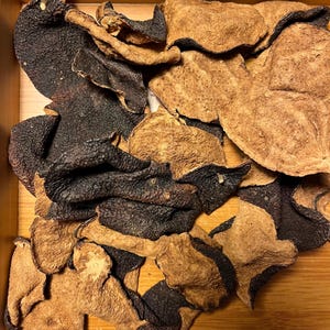 Pode incluir: Close-up de cascas de citrinos secas, com uma mistura de pedaços castanhos escuros e castanhos claros. As cascas têm uma aparência texturizada e enrugada, e estão dispostas num recipiente de madeira. A imagem destaca as cores e texturas naturais da fruta seca.