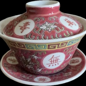 Puede incluir: Un juego de té gaiwan de cerámica que incluye un cuenco, una tapa y un plato en color rojo intenso con intrincados patrones florales y medallones circulares blancos con caracteres chinos rojos. El borde presenta una cenefa geométrica.
