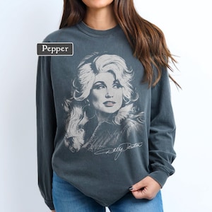 Op de afbeelding: Een antracietgrijs sweatshirt met lange mouwen en een vervaagde witte grafische print van Dolly Parton met haar handtekening eronder. Een klein rechthoekig label in de linkerbovenhoek bevat de tekst Pepper in witte letters.