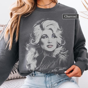 Puede incluir: Una sudadera de cuello redondo y manga larga en color gris carbón con un gran estampado gráfico blanco detallado de Dolly Parton, mostrando su característico cabello voluminoso y pendientes de aro. El texto "Charcoal" aparece en un recuadro arriba.