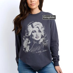 Op de afbeelding: Een antracietgrijs sweatshirt met lange mouwen en een ronde hals, voorzien van een witte grafische illustratie van Dolly Parton met volumineus haar. Het woord Graphite staat in een klein wit kader rechtsboven en de handtekening Dolly Parton staat in wit cursief schrift onder het portret.