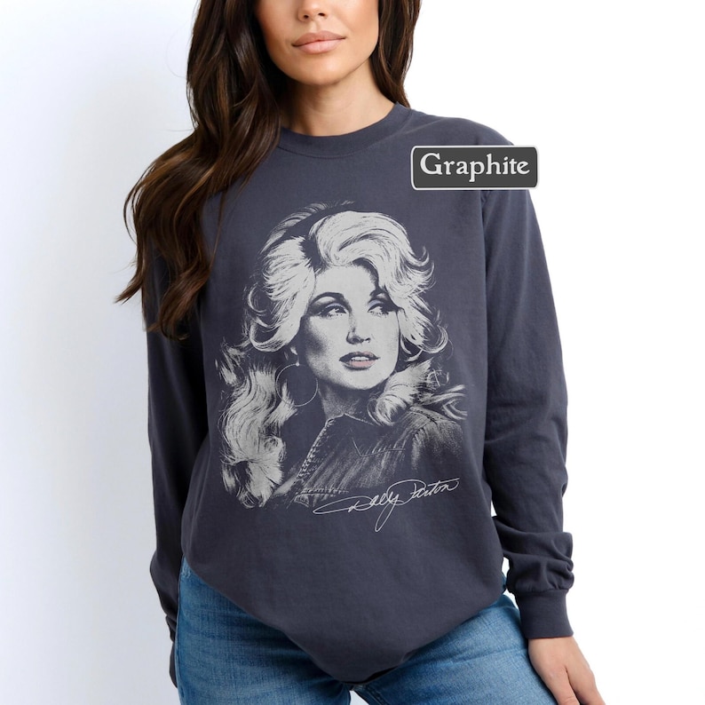 Op de afbeelding: Een antracietgrijs sweatshirt met lange mouwen en een ronde hals, voorzien van een witte grafische illustratie van Dolly Parton met volumineus haar. Het woord Graphite staat in een klein wit kader rechtsboven en de handtekening Dolly Parton staat in wit cursief schrift onder het portret.
