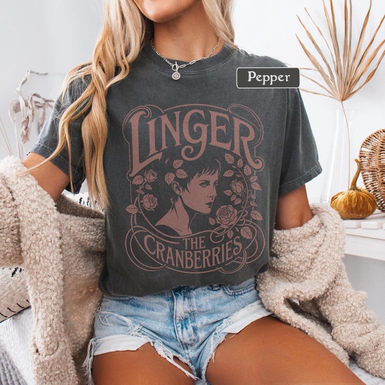 May include: Dark gray t-shirt featuring the text "Linger" above an illustration of a woman's profile, surrounded by roses, with "The Cranberries" below. The shirt is a vintage style, with a slightly faded appearance.