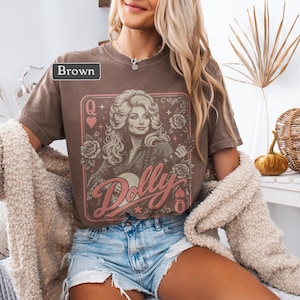 May include: A brown short-sleeve t-shirt featuring a graphic print of a woman with long blonde hair, roses, and the text "Dolly" in a pink script font. The design is framed like a playing card with a "Q" and heart symbols in the corners.