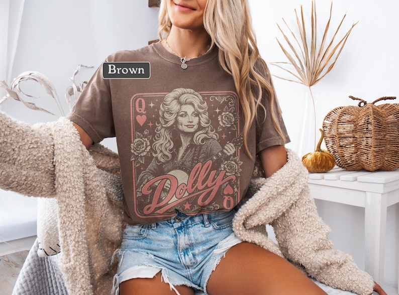 May include: A brown short-sleeve t-shirt featuring a graphic print of a woman with long blonde hair, roses, and the text "Dolly" in a pink script font. The design is framed like a playing card with a "Q" and heart symbols in the corners.