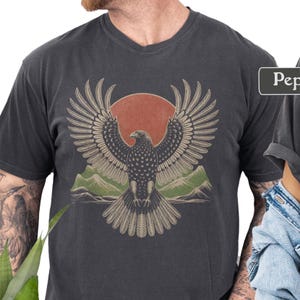 May include: Dark gray v-neck t-shirt featuring a detailed graphic of an eagle with outstretched wings, a red sun, and green mountains. The eagle design is centered on the shirt.