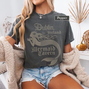 May include: Dark grey t-shirt featuring the text "Dublin, Ireland Mermaid Tavern" and a mermaid illustration. The t-shirt is a crew neck with short sleeves. The outfit includes denim shorts and a cream-coloured cardigan.