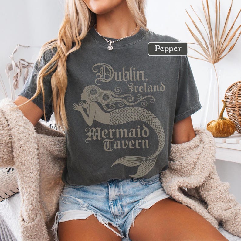 May include: Dark grey t-shirt featuring the text "Dublin, Ireland Mermaid Tavern" and a mermaid illustration. The t-shirt is a crew neck with short sleeves. The outfit includes denim shorts and a cream-coloured cardigan.