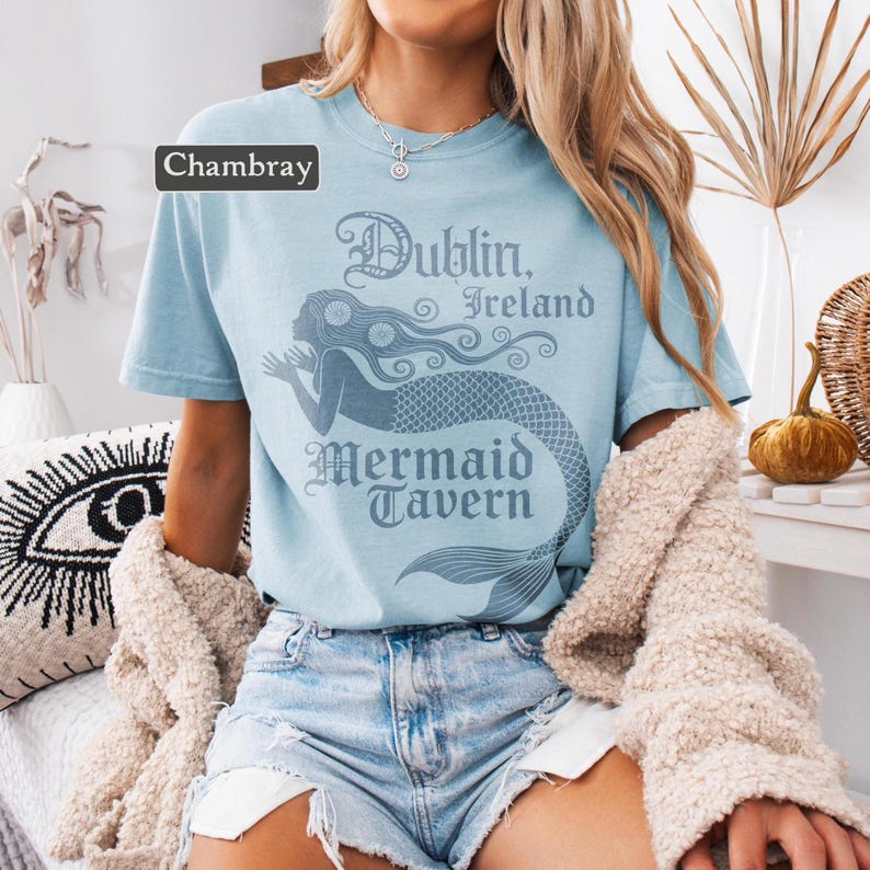 May include: Pale blue t-shirt featuring a mermaid design and the words "Dublin, Ireland Mermaid Tavern". The top is a short-sleeved, crew neck style.