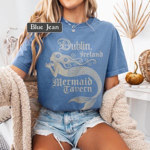 May include: Light blue t-shirt with the text "Dublin, Ireland Mermaid Tavern" and a mermaid graphic in a light beige colour. The shirt is a blue jean colour.