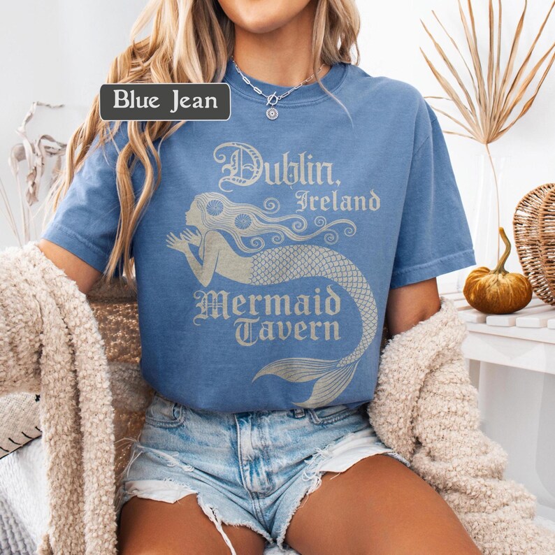 May include: Light blue t-shirt with the text "Dublin, Ireland Mermaid Tavern" and a mermaid graphic in a light beige colour. The shirt is a blue jean colour.