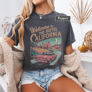 May include: Dark gray t-shirt with a vintage-style graphic that reads "Welcome to The Hotel California." The design features a pink convertible car on a winding road, a building, and a crescent moon. The shirt is a relaxed fit.