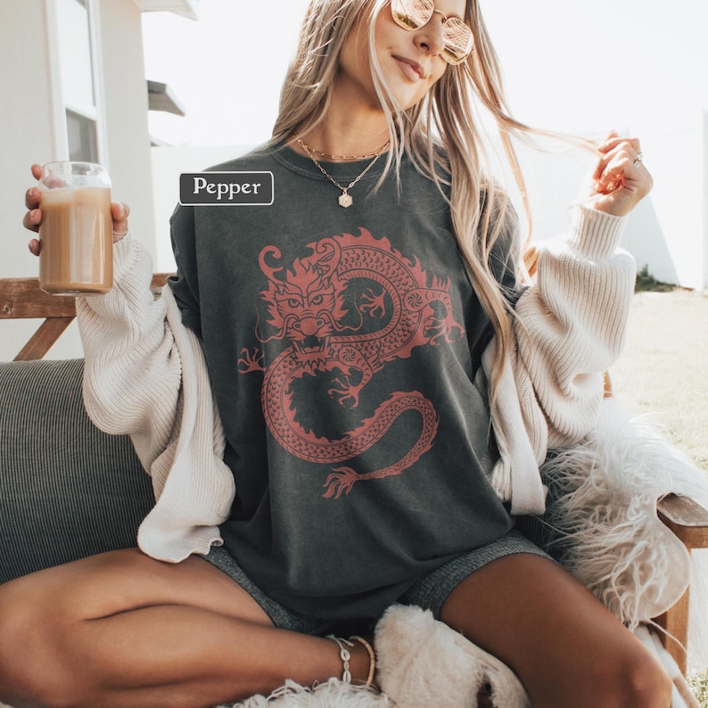 May include: Dark gray long-sleeve shirt with a red dragon graphic. The word "Pepper" is in a white box on the upper left. The shirt is worn with gray shorts and a cream-colored cardigan. A glass of iced coffee is held in the left hand.