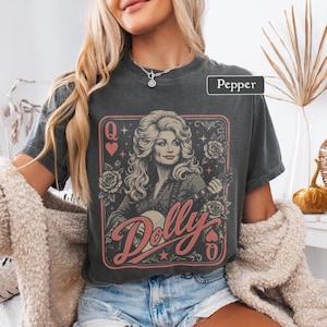 May include: A charcoal gray short-sleeve t-shirt featuring a graphic print of a woman with voluminous hair playing a guitar. The design includes roses, stars, heart symbols, the letter Q, and the word Dolly in a script font. The text Pepper is visible.